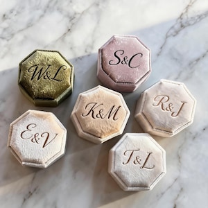 May include: Six octagonal velvet ring boxes in various colors including olive green, dusty rose, and cream. Each box features a custom monogram engraved on the lid, specifically the letters W&L, S&C, K&M, R&J, E&V, and T&L in a classic serif font.