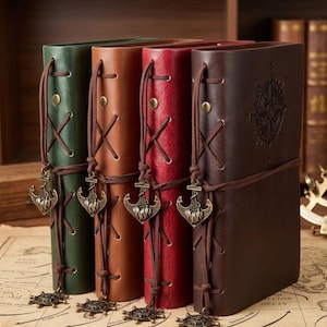 May include: Four vertical journals in green, tan, red, and dark brown. Each features a leather-like cover, side lacing with brown cord, a brass-toned anchor charm, and a metal ship wheel charm at the base. The dark brown journal has a compass design.