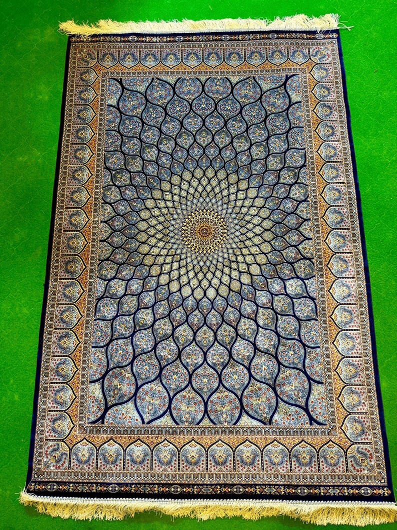 Puede incluir: Una alfombra rectangular que presenta un medall&oacute;n central con un patr&oacute;n floral radiante en tonos azul, crema y dorado. La alfombra tiene un borde azul oscuro con motivos repetitivos intrincados y flecos claros en los extremos superior e inferior.
