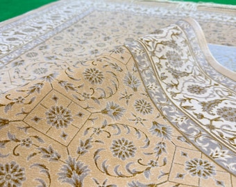 Puede incluir: Una alfombra rectangular con fondo beige claro y complejos patrones florales y de enredaderas en tonos blanco y gris. La alfombra presenta un borde detallado con formas geométricas y motivos florales, rematado con flecos en el extremo.