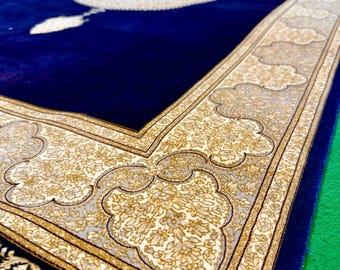 Puede incluir: Una alfombra de oración rectangular que presenta un centro azul profundo con un medallón circular central y un motivo de arco apuntado. El borde ancho muestra intrincados patrones florales en crema y oro, con flecos blancos.