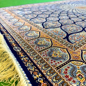 Puede incluir: Una alfombra rectangular que presenta un campo azul profundo con un patr&oacute;n repetitivo de motivos florales en azul claro y dorado. El borde muestra intrincados dise&ntilde;os bot&aacute;nicos en tonos rojos, dorados y azules, terminados con largos flecos color crema.