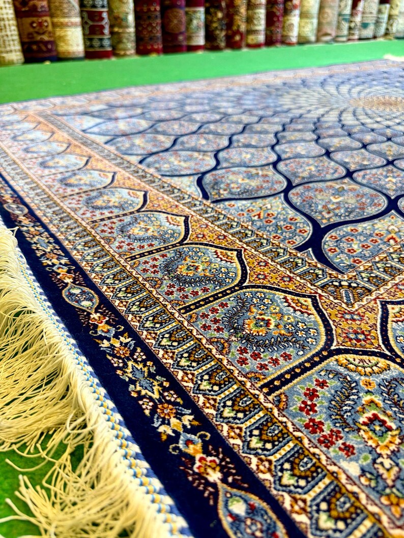 Puede incluir: Una alfombra rectangular que presenta un campo azul profundo con un patr&oacute;n repetitivo de motivos florales en azul claro y dorado. El borde muestra intrincados dise&ntilde;os bot&aacute;nicos en tonos rojos, dorados y azules, terminados con largos flecos color crema.