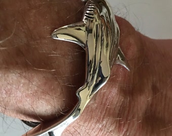 May include: A silver shark ring. The ring is highly polished, reflecting light. The shark design features detailed fins and a textured body. The ring is worn on a wrist, showcasing the jewelry's design and craftsmanship. The ring is a statement piece.