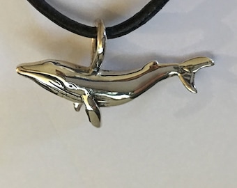 Op de afbeelding: Een zilveren walvis hanger aan een zwart koord. De hanger is een gedetailleerde, driedimensionale sculptuur van een walvis, met vinnen en een staart. Het koord is een eenvoudig, rond, zwart materiaal, waarschijnlijk leer. De achtergrond is een effen wit oppervlak.