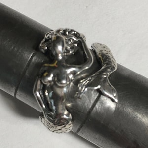May include: A silver ring featuring a mermaid design. The ring depicts a mermaid with flowing hair and a fishtail, wrapped around the band. The ring is shown on a gray metal ring sizer.