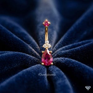 May include: A gold curved barbell belly ring featuring a round-cut red gemstone at the top, a cluster of three small clear stones in the center, and a pear-shaped red gemstone at the bottom. The text Knovajewels is visible below the jewelry.