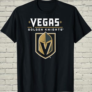 May include: A black short-sleeve t-shirt featuring the white text VEGAS and GOLDEN KNIGHTS with gold star accents. Below the text is a shield-shaped logo displaying a stylized knight helmet in gold, grey, and white, with a V-shaped cutout in the center.