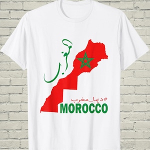 May include: A white short-sleeved t-shirt featuring a red map of Morocco with a green star in the center. The design includes green Arabic calligraphy, the red Arabic text #ديما_مغرب, and the word MOROCCO printed in large green capital letters.