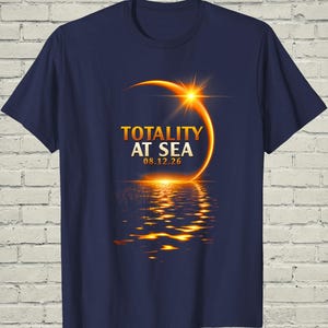 May include: A navy blue short-sleeve t-shirt featuring a graphic of a solar eclipse over water. The text reads TOTALITY AT SEA 08.12.26 in gold lettering. The design includes a bright sun flare and a golden reflection shimmering on the ocean surface.