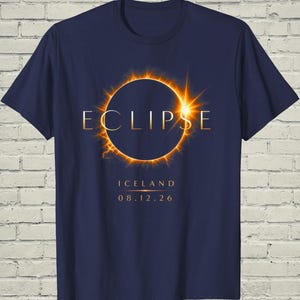 May include: A navy blue short-sleeve t-shirt featuring a graphic of a solar eclipse with a glowing orange corona. The word ECLIPSE is printed in gold, with ICELAND and the date 08.12.26 positioned below the celestial event in gold lettering.