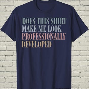 May include: A navy blue short-sleeve t-shirt featuring the text "DOES THIS SHIRT MAKE ME LOOK PROFESSIONALLY DEVELOPED" printed in a stacked layout. The words "DOES THIS SHIRT MAKE ME LOOK" are light blue, "PROFESSIONALLY" is pink, and "DEVELOPED" is tan.