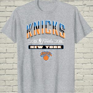 May include: A heather gray short-sleeve t-shirt featuring the text KNICKS in blue, black, and orange. Below, the text 20 FINALS 26 surrounds an NBA logo. A black rectangular box contains the text NEW YORK, and a New York Knicks logo is below.