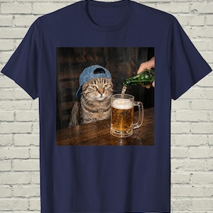 May include: A navy blue t-shirt featuring a graphic print of a brown tabby cat wearing a blue denim baseball cap. The cat is positioned next to a glass beer mug being filled with golden liquid from a green glass bottle, set against a dark wood background.