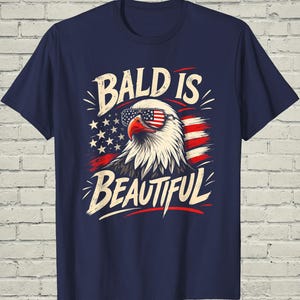 May include: A navy blue short-sleeve t-shirt featuring a graphic of a bald eagle wearing sunglasses with an American flag pattern. The eagle is set against a stylized flag background with the text BALD IS BEAUTIFUL in a bold, cream-colored font.