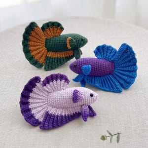Peut inclure: Trois jouets poissons combattants crochetés à la main. L'un est vert foncé avec des accents orange, un est violet profond avec des nageoires bleu vif, et un est lavande clair avec des bords de nageoires violet foncé. Yeux en perles noires.