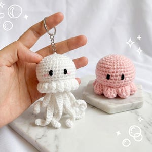 May include: A white crocheted jellyfish keychain with black eyes and pink cheeks, featuring a metal ring and chain. Beside it sits a small, round, light pink crocheted octopus plush toy with black eyes, resting on a white marble-patterned base.