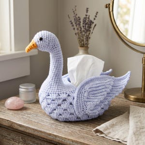 May include: A light purple crochet tissue box cover shaped like a swan with a textured body, a long neck, and an orange beak. The swan features detailed wing patterns and holds a white tissue upright within its back, displaying a handcrafted design.