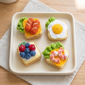 May include: Four square crochet toast pieces topped with various food items. One has two orange sausages and green lettuce, one has a fried egg and lettuce, one has blue and red berries, and one has pink shrimp and green lettuce on a square tray.