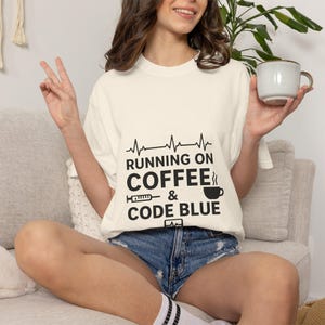 Könnte beinhalten: Cremefarbenes T-Shirt mit dem schwarzen Text "Running on Coffee & Code Blue". Das Design umfasst eine Herzschlaglinie, eine Kaffeetasse und eine Spritze. Die Person trägt Jeansshorts und hält eine Kaffeetasse.