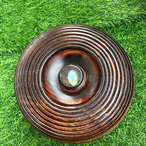 May include: A round, wooden bowl with a dark brown finish. The bowl features concentric circular grooves and a small, central knob. A sticker with the text "BEST QUALITY" is visible in the center. The bowl is set against a green background.