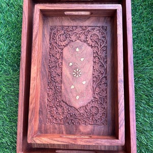 May include: Set of two wooden serving trays. The trays are rectangular with carved floral designs and integrated handles. The wood has a rich, reddish-brown color. The inner tray features a detailed floral carving with gold accents.