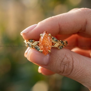 May include: A gold ring featuring a marquise-cut, orange sunstone gemstone. The band is adorned with leaf and small green gemstone accents. The ring is held up against a blurred background.