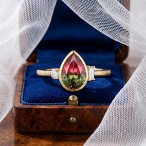 May include: A gold ring featuring a central pear-shaped bicolor tourmaline gemstone that transitions from pink to green. The center stone is bezel-set and flanked by a small round brilliant-cut diamond on one side and a baguette-cut diamond on the other.