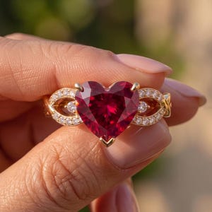May include: A gold ring featuring a large, faceted, heart-shaped red gemstone at the centre. The band is crafted with an intricate, openwork design on both sides, embellished with small, round, clear white stones set along the metal frame.