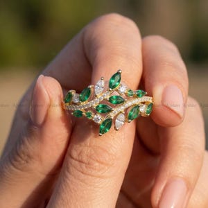 May include: A gold ring featuring a leaf design with emerald green and white gemstones. The ring has a delicate, nature-inspired aesthetic, with the gemstones set along the band in a leaf-like pattern. The ring is held by a person.