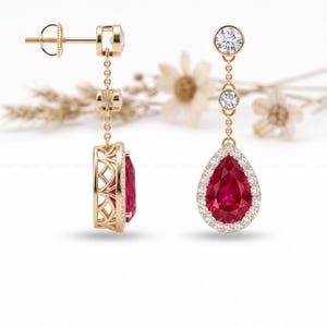 May include: A pair of gold-colored drop earrings. Each earring features a large, pear-shaped, red gemstone surrounded by small, clear stones. The earrings have a delicate chain design with additional clear stones and a post-and-stud closure.