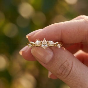 May include: A gold ring with a delicate design, featuring a central pear-shaped stone and smaller stones. The ring's band has a woven pattern, and the stones sparkle. The ring is held in a hand against a blurred green background.