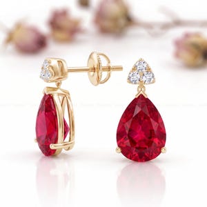 Può includere: Orecchini pendenti in oro con pietre preziose rosse a forma di pera. Ogni orecchino ha un piccolo triangolo di pietre bianche nella parte superiore e un perno d'oro. Sfondo sfocato.