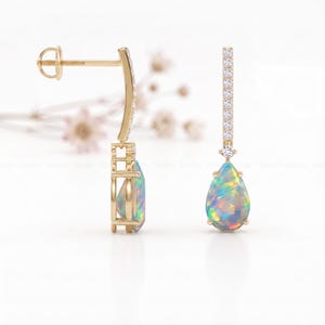 May include: A pair of gold-colored drop earrings. Each earring features a pear-shaped opal gemstone with iridescent colors. The earrings have a post-back closure and are accented with small, clear gemstones.