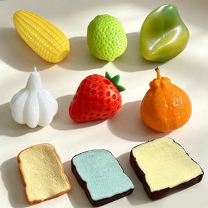 May include: A collection of nine food-shaped items including a yellow corn cob, a textured lime, a smooth green fruit, a white garlic bulb, a red strawberry, an orange citrus fruit, and three slices of toast with different colored toppings.