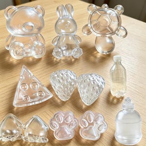 Könnte beinhalten: Eine Sammlung durchsichtiger Kunststoffformen in verschiedenen Ausführungen, darunter ein Bär, ein Hase, eine Schildkröte, ein Dreieck, zwei Erdbeeren, zwei Pfotenabdrücke, zwei kleine abgerundete Figuren, eine Flasche und ein runder Behälter.