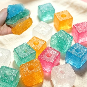 May include: A collection of translucent, cube-shaped beads with a textured, crystalline surface. Each bead features a central circular hole. The cubes are available in various colors including blue, green, orange, pink, and clear white.