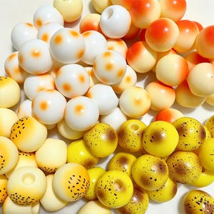 May include: A collection of spherical beads featuring a mix of solid white beads with orange centers, solid yellow beads, and yellow beads with brown speckled patterns. Each bead has a central hole for threading, creating a textured, multi-colored assortment.