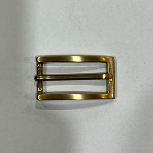 Può includere: Una fibbia per cintura rettangolare con finitura in oro spazzolato. La fibbia presenta un design a cornice semplice con una barra orizzontale centrale e un ardiglione mobile fissato su un lato, creando un componente hardware funzionale.