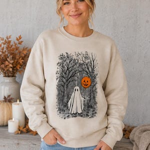 Könnte beinhalten: Ein beigefarbenes Sweatshirt mit Rundhalsausschnitt und einem Grafikdruck eines weißen Geistes, der in einem dunklen Wald steht und einen orangefarbenen Kürbisballon hält. Die Illustration ist in einem detaillierten Skizzenstil gehalten.