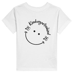 Könnte beinhalten: Ein weißes T-Shirt mit kurzen Ärmeln und einem schwarzen Grafikdruck auf der Brust. Das Design zeigt ein Smiley-Gesicht mit zwei Punkten als Augen und einer geschwungenen Linie als Mund, wobei der Text Kindergartenkind 2026 integriert ist.