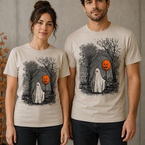 Könnte beinhalten: Ein beiges Kurzarm-T-Shirt mit Rundhalsausschnitt und einer schwarz-weißen Tuschezeichnung eines Geistes in einem Wald, der einen leuchtend orangefarbenen Kürbis-Luftballon hält. Die Grafik befindet sich mittig auf der Brust.