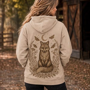 Könnte beinhalten: Ein beigefarbener Kapuzenpullover mit einer detaillierten braunen Grafik auf der Rückseite. Das Design zeigt einen zentralen Fuchs, der inmitten eines Blumenkranzes sitzt, umgeben von vier Motten und einem Halbmond über dem Kopf des Fuchses.