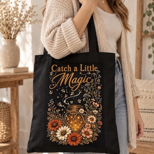 Könnte beinhalten: Eine schwarze Tragetasche mit einem Grafikdruck eines mit leuchtenden Lichtern gefüllten Glases, umgeben von orangefarbenen, weißen und braunen Blumen und Glühwürmchen. Der Text "Catch a Little Magic" steht in goldener Schrift über dem Glas.