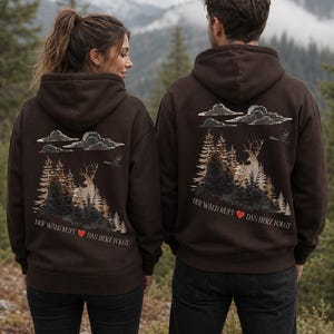 Könnte beinhalten: Ein dunkelbrauner Kapuzenpullover mit einem Grafikdruck auf der Rückseite. Das Motiv zeigt eine Waldszene mit Tannen, einem Hirsch, Wolken und einem Vogel. Unter der Illustration steht der Text: DER WALD RUFT ❤️ DAS HERZ FOLGT.