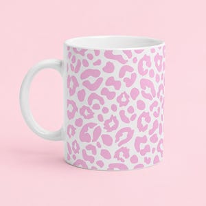 Könnte beinhalten: Ein weißer Keramikbecher mit einem sich wiederholenden rosa Leopardenmuster auf der Außenfläche. Der Becher hat einen geschwungenen Henkel auf der linken Seite und einen glatten, zylindrischen Körper mit einem sauberen weißen Innenrand.