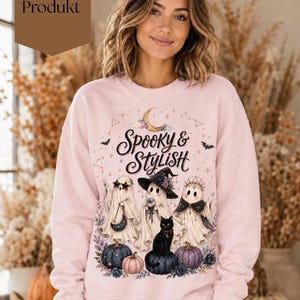 Könnte beinhalten: Ein hellrosa Sweatshirt mit Rundhalsausschnitt und einem Grafikdruck von drei Geistern, einer schwarzen Katze, Kürbissen und einer Mondsichel. Der Text "Spooky & Stylish" ist mittig über den Geistern gedruckt, umgeben von Sternen und kleinen Fledermäusen.