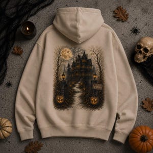 Könnte beinhalten: Ein beigefarbener Kapuzenpullover mit einer detaillierten Grafik auf der Rückseite. Der Druck zeigt ein dunkles, mehrstöckiges Herrenhaus bei Nacht unter einem Vollmond, mit einem Steinweg zum Eingang, zwei leuchtenden Kürbissen und einem Schild Haunted Villa.