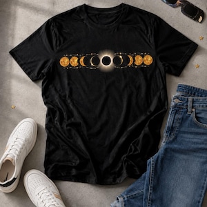 Könnte beinhalten: Ein schwarzes Kurzarm-T-Shirt mit einem horizontalen Grafikdruck der Mondphasen in Gold- und Gelbtönen, der mittig auf der Brust platziert ist. Das Design zeigt eine Abfolge von Mondzyklen, beginnend bei einer Mondsichel bis hin zur totalen Finsternis.