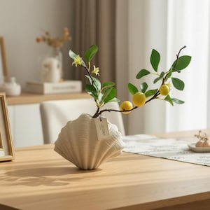 Può includere: Un vaso beige a forma di conchiglia contiene un ramo con foglie verdi e frutti gialli. Il vaso è appoggiato su un tavolo di legno. Una piccola etichetta pende dal ramo. Lo sfondo comprende una finestra e altri oggetti decorativi.